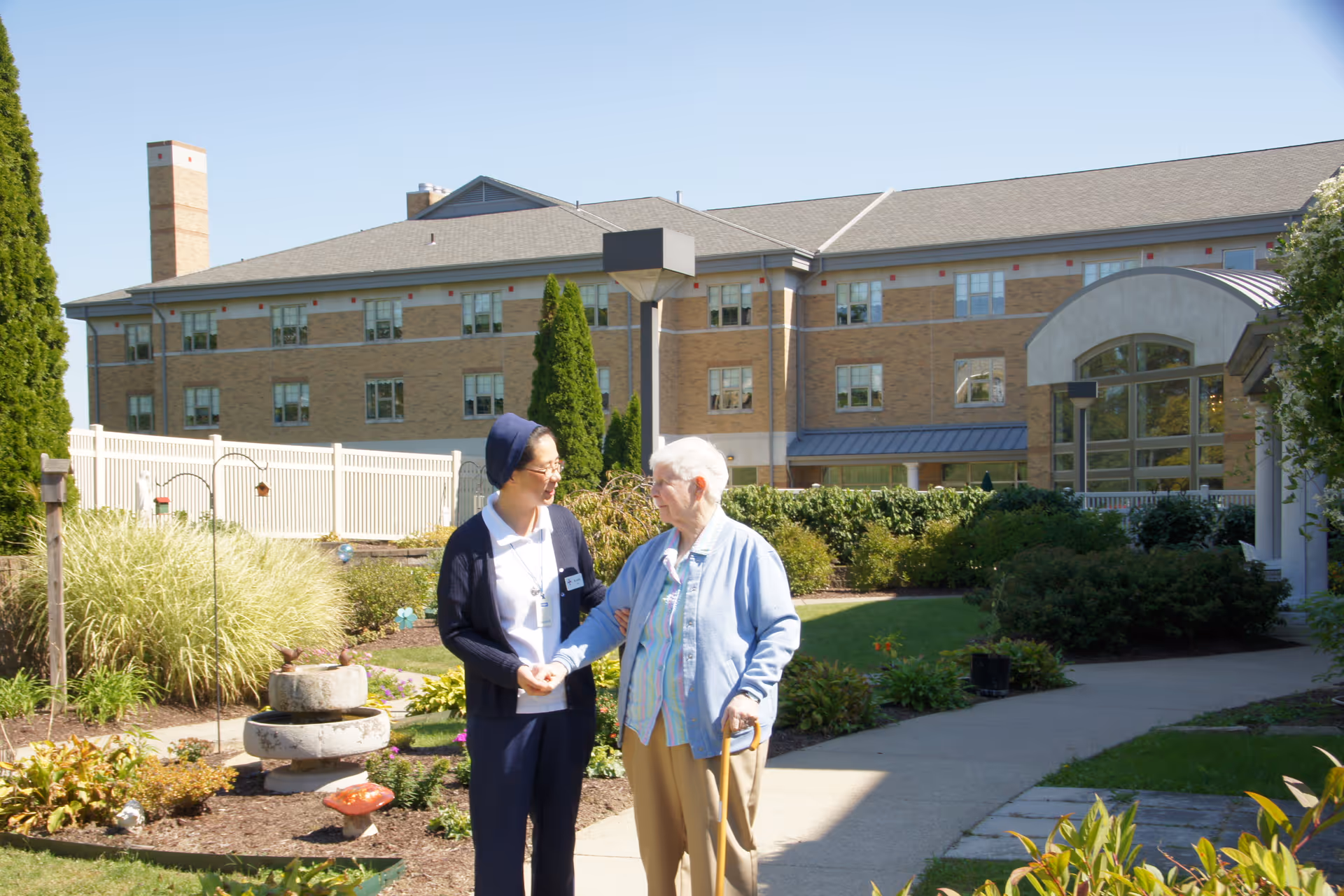 An elderly woman with a cane is walking outside in a garden area of a senior living facility, accompanied by a caregiver. They are holding hands and smiling at each other. In the background, there is a large brick building with multiple windows and a white fence surrounding the garden.