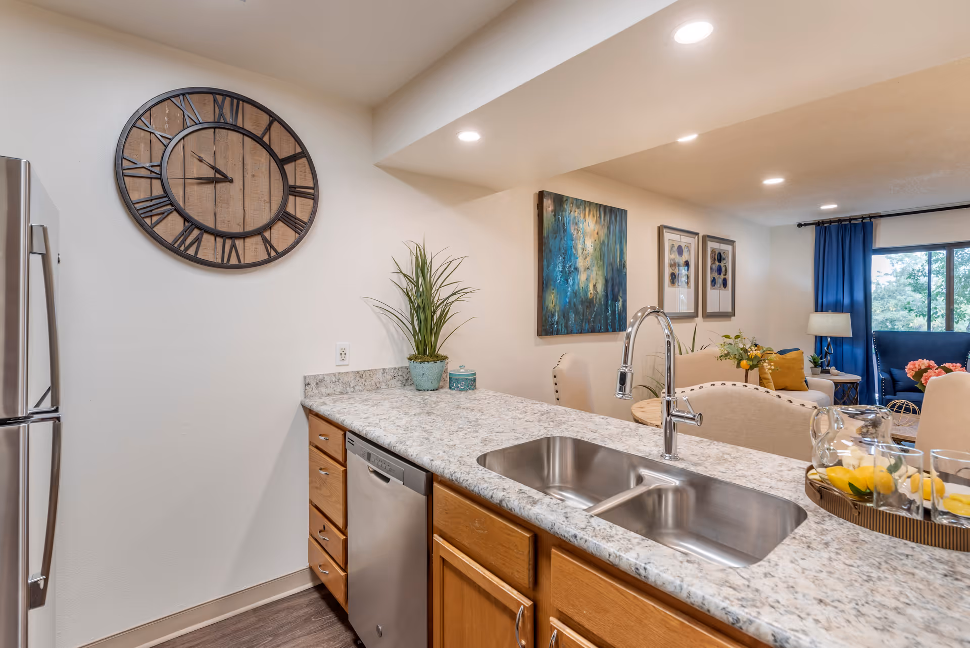 Interior view of a kitchen area with a granite countertop, stainless steel sink and faucet, wooden cabinets, and a stainless steel dishwasher. A large decorative wall clock is mounted on the wall. In the background, a living room area is visible with beige chairs, a blue armchair, framed artwork on the walls, a lamp, and a window with blue curtains.