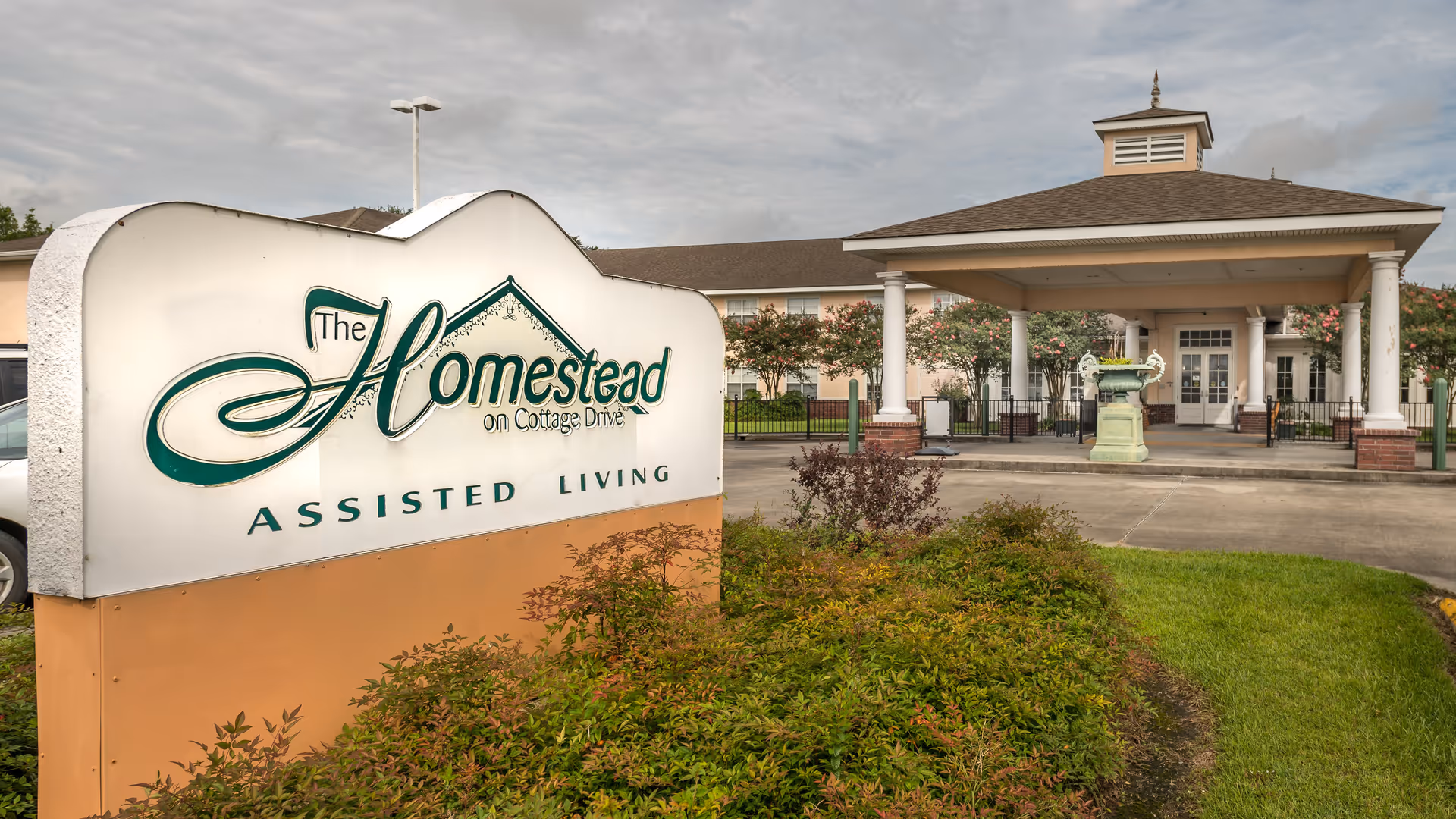 Entrance view of The Homestead Assisted Living facility showing a large sign with the facility's name and a covered drop-off area with columns in front of the building, surrounded by greenery and a cloudy sky.