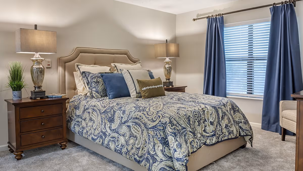 A neatly made bed with a beige upholstered headboard and a blue and beige paisley patterned comforter. The bed has multiple pillows in shades of blue, beige, and green. On either side of the bed are wooden nightstands with matching lamps and a small plant on the left nightstand. A window with blue curtains and blinds is on the right side of the room, and a beige armchair is partially visible in the corner.