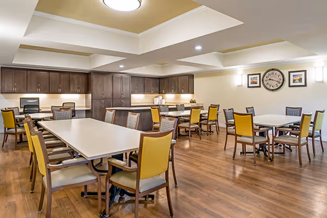 A spacious dining room with multiple tables and chairs arranged neatly. The room features wooden flooring, a tray ceiling with recessed lighting, and dark wood cabinetry along the back wall. The walls are decorated with framed pictures and a large clock.