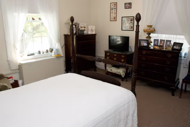 A bedroom with a large wooden four-poster bed covered with a white bedspread. There are two windows with white curtains, a wooden dresser with a TV on top, another dresser with framed photos and a vintage lamp, and a chair in the corner.