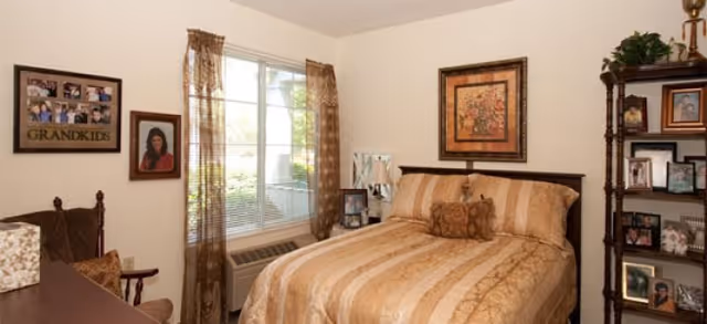 A cozy bedroom with a neatly made bed featuring a beige and gold striped comforter and matching pillows. There is a window with sheer brown curtains allowing natural light to enter. On the wall above the bed hangs a framed floral artwork. To the right of the bed is a dark wooden shelving unit displaying framed photos and decorative items. On the left side, there is a rocking chair and framed pictures on the wall, including one labeled 'GRANDKIDS'.