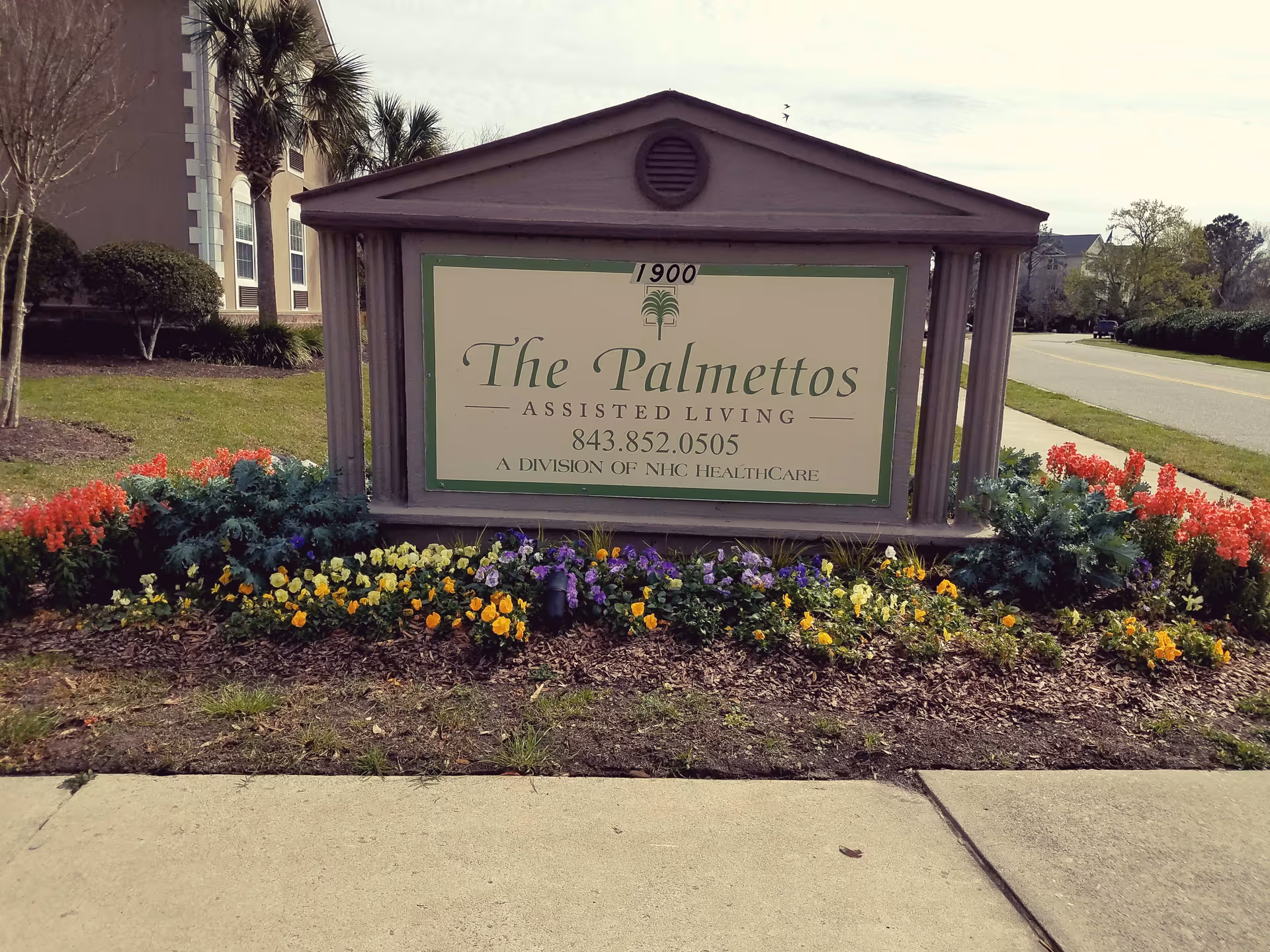 Outdoor sign for The Palmettos Assisted Living, surrounded by colorful flowers and greenery, with a building and road in the background.