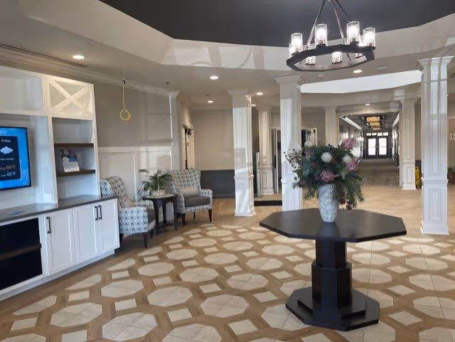 A spacious and well-lit interior common area with a patterned floor, white pillars, and a dark ceiling. There is a black octagonal table in the center with a floral arrangement. To the left, there are two upholstered chairs with a small table between them, and built-in white cabinetry with a TV mounted above. The hallway extends into the background with more lighting fixtures and doors.