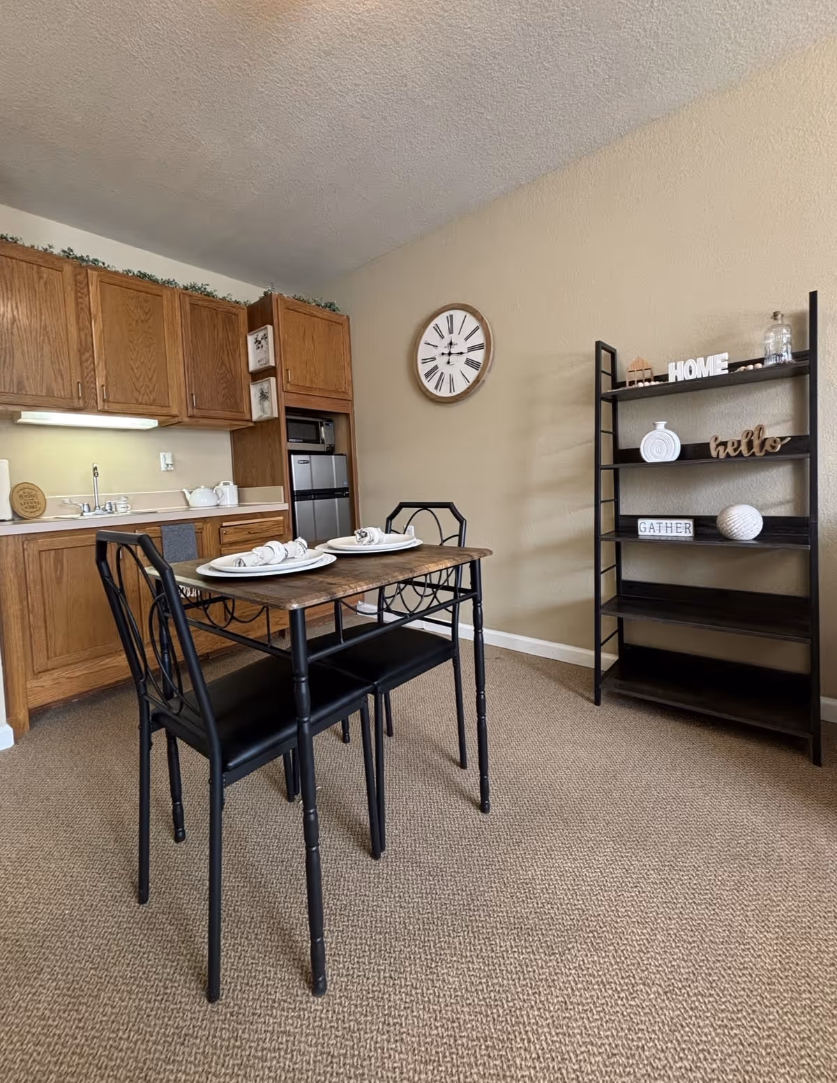 A small dining area with a wooden table set for two with plates and napkins, two black metal chairs, a wooden kitchen with cabinets and a sink in the background, a wall clock, and a black shelving unit with decorative items including signs that say HOME, hello, and GATHER.