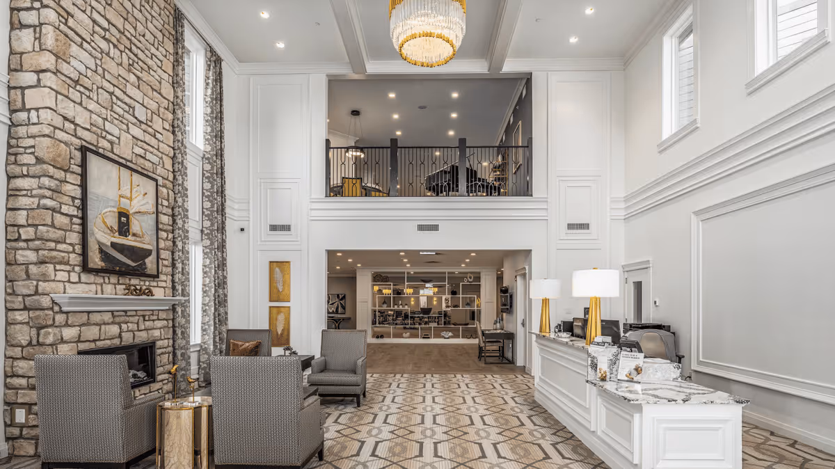 Spacious and elegant senior living facility common area with high ceilings, a large stone fireplace on the left, comfortable seating with armchairs and a small table, a reception desk with two lamps on the right, and a mezzanine level with a piano and additional seating above.