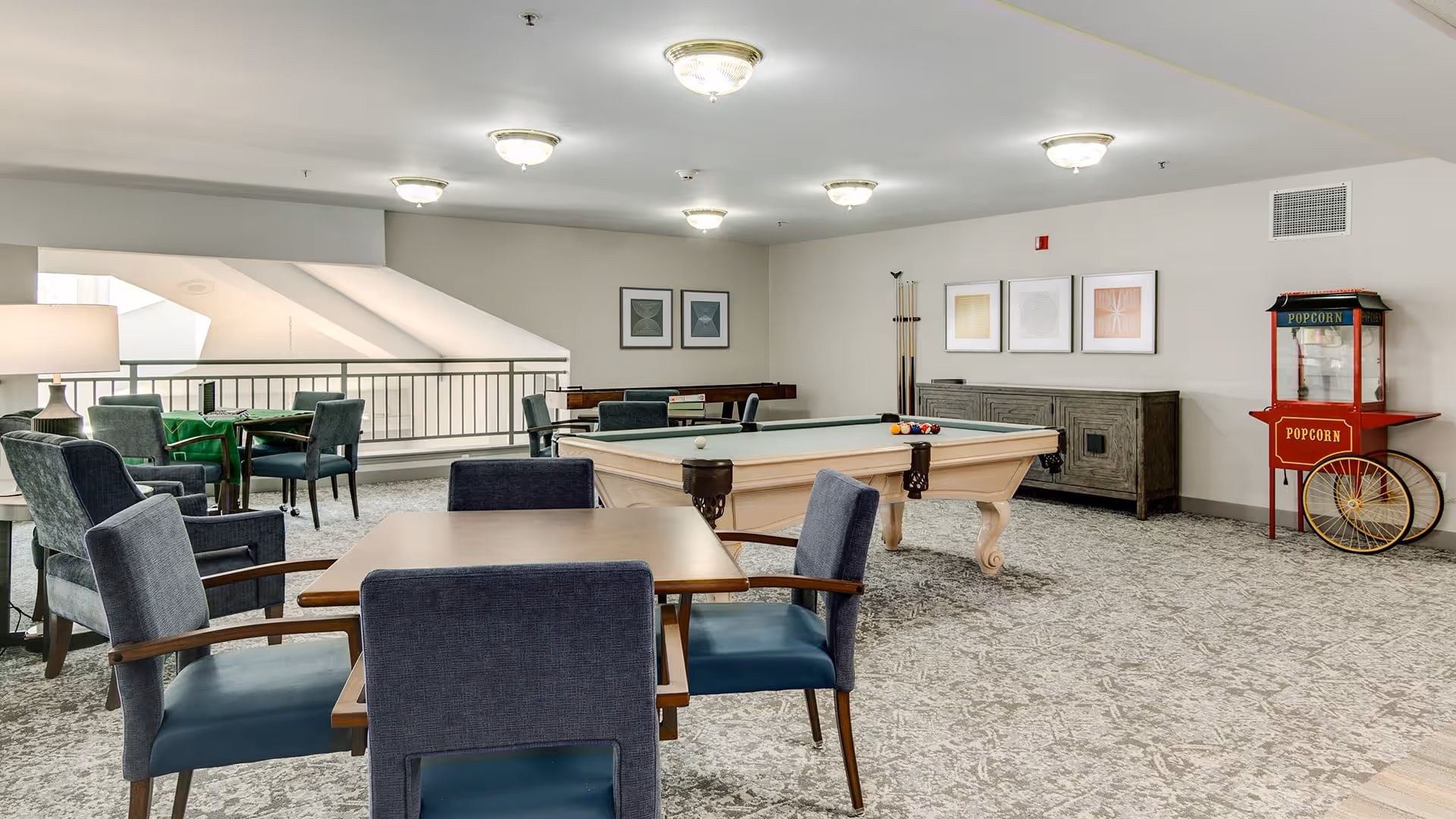 A spacious recreational room with a pool table, several tables with chairs, a popcorn machine, and framed artwork on the walls. The room has carpeted flooring and ceiling lights, with a railing overlooking a lower level.