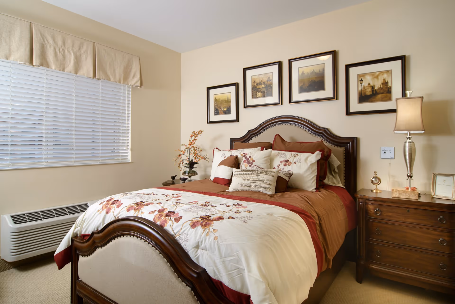 A neatly made bed with floral patterned bedding and multiple pillows in a bedroom. The room has beige walls with four framed pictures hanging above the bed. There are two wooden nightstands on either side of the bed, one with a lamp and decorative items, and the other with a small plant. A window with closed blinds and a beige valance is on the left side of the room.