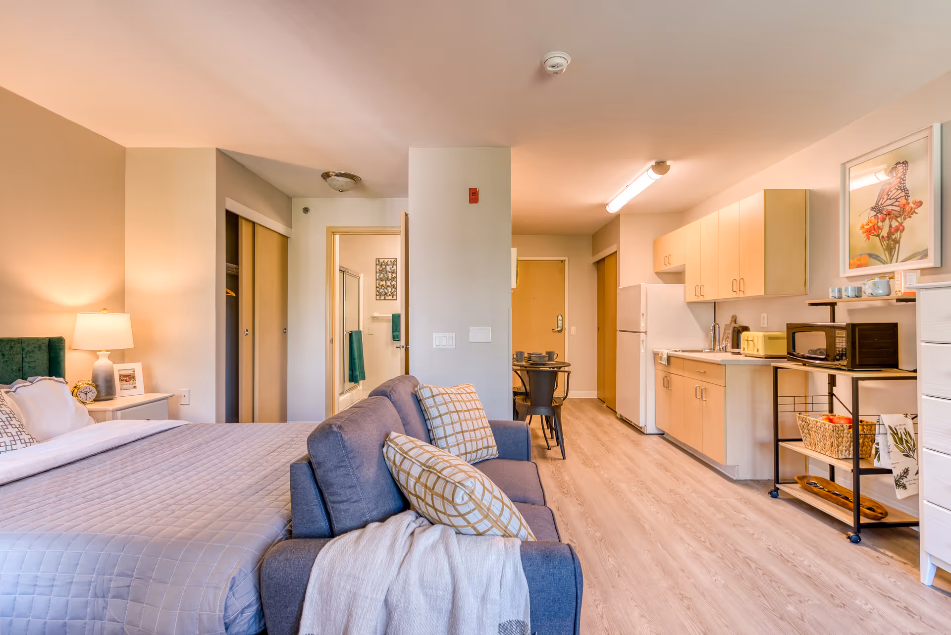 A bright and cozy studio apartment in a senior living facility featuring a bed with a green headboard and gray bedding, a gray couch with patterned pillows, a small dining table with two chairs, and a kitchenette with light wood cabinets, a refrigerator, microwave, and toaster. The room has light wood flooring and a bathroom visible through an open door.