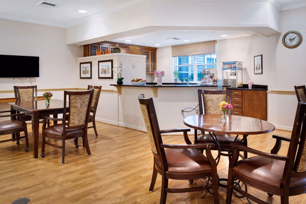 Bright communal dining area with wooden tables and chairs, a service counter and wall-mounted TV.