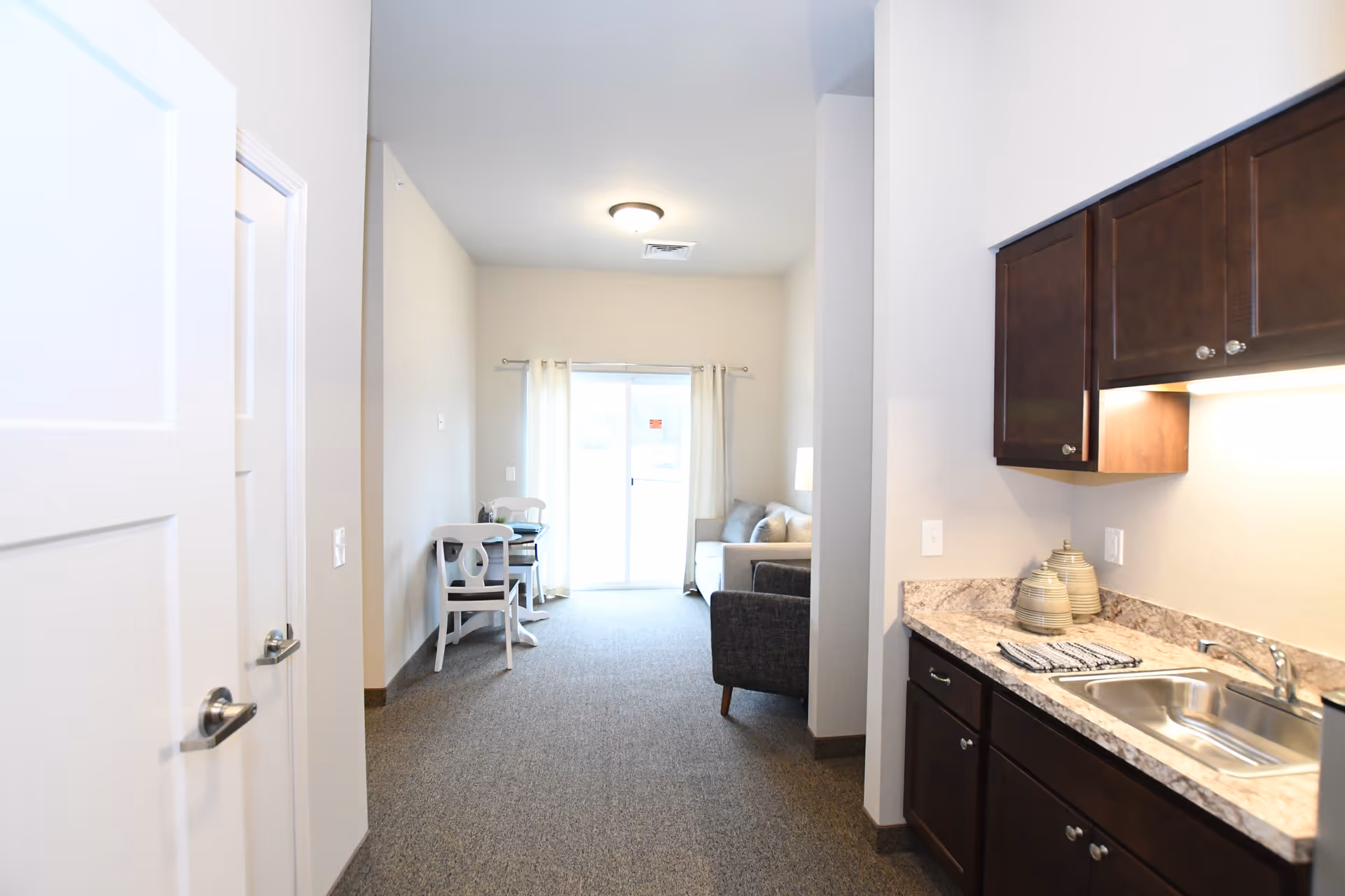 Interior view of an assisted living apartment showing a kitchenette with dark cabinets and sink on the right and a small dining and living area with sliding glass door ahead.