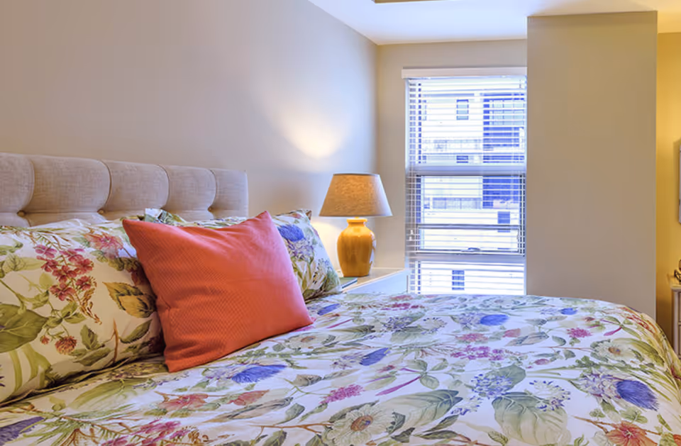 A bedroom with a bed featuring floral-patterned bedding and a large coral-colored pillow. There is a beige upholstered headboard and a bedside table with a yellow lamp. A window with white blinds is visible in the background.