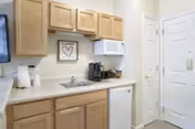 Small kitchenette area with light wood cabinets, a white microwave, a coffee maker, a small sink, a mini refrigerator, and a framed artwork on the wall. There is a door to the right side and a wall-mounted TV partially visible on the left.