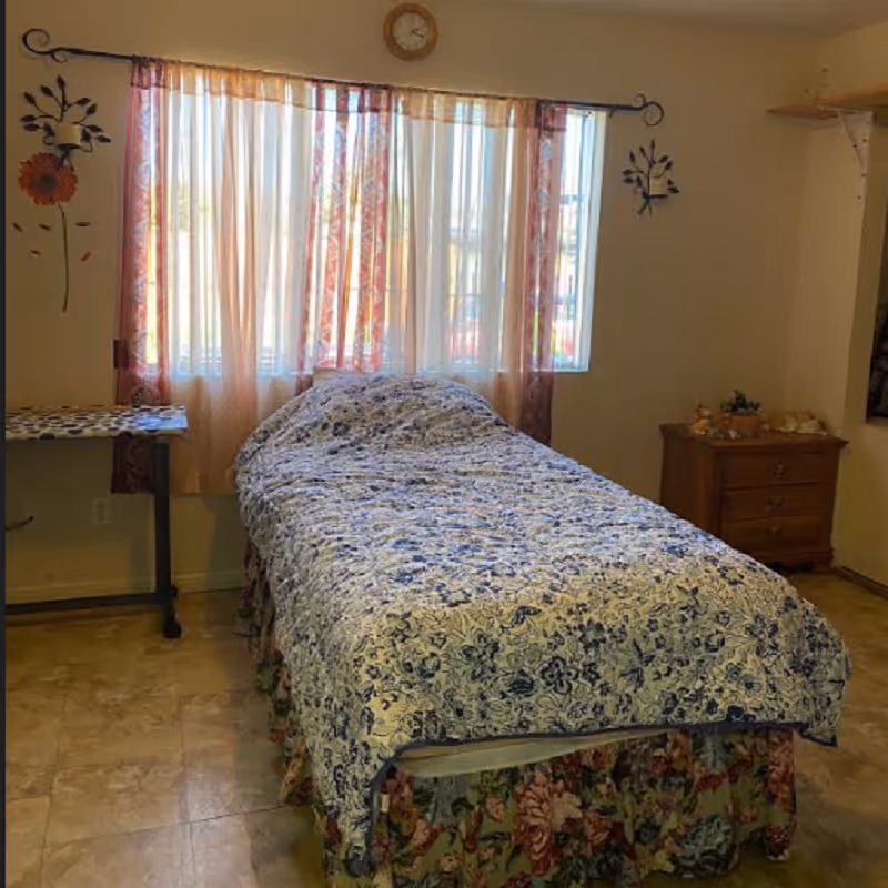 A single bed with floral quilt in a small bedroom with curtains and a dresser.