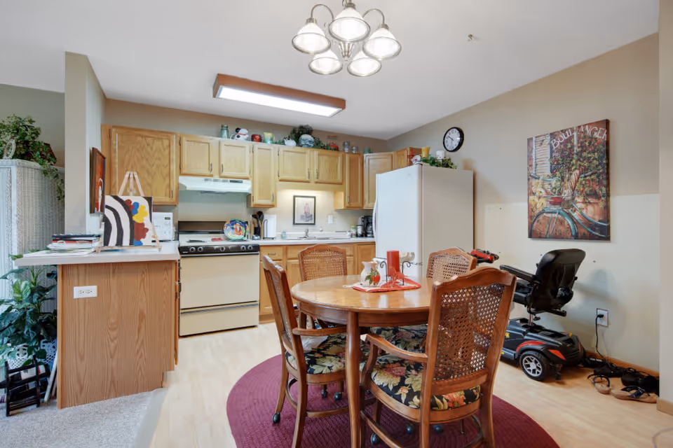A cozy kitchen and dining area in a senior living facility. The kitchen features wooden cabinets, a white refrigerator, a stove with an overhead vent, and various kitchen items on the counters. In front of the kitchen is a round wooden dining table with four chairs that have floral cushions. A red circular rug is underneath the table. On the right side, there is a mobility scooter parked against the wall, which is decorated with a colorful painting and a clock. The room is well-lit with a ceiling light fixture.