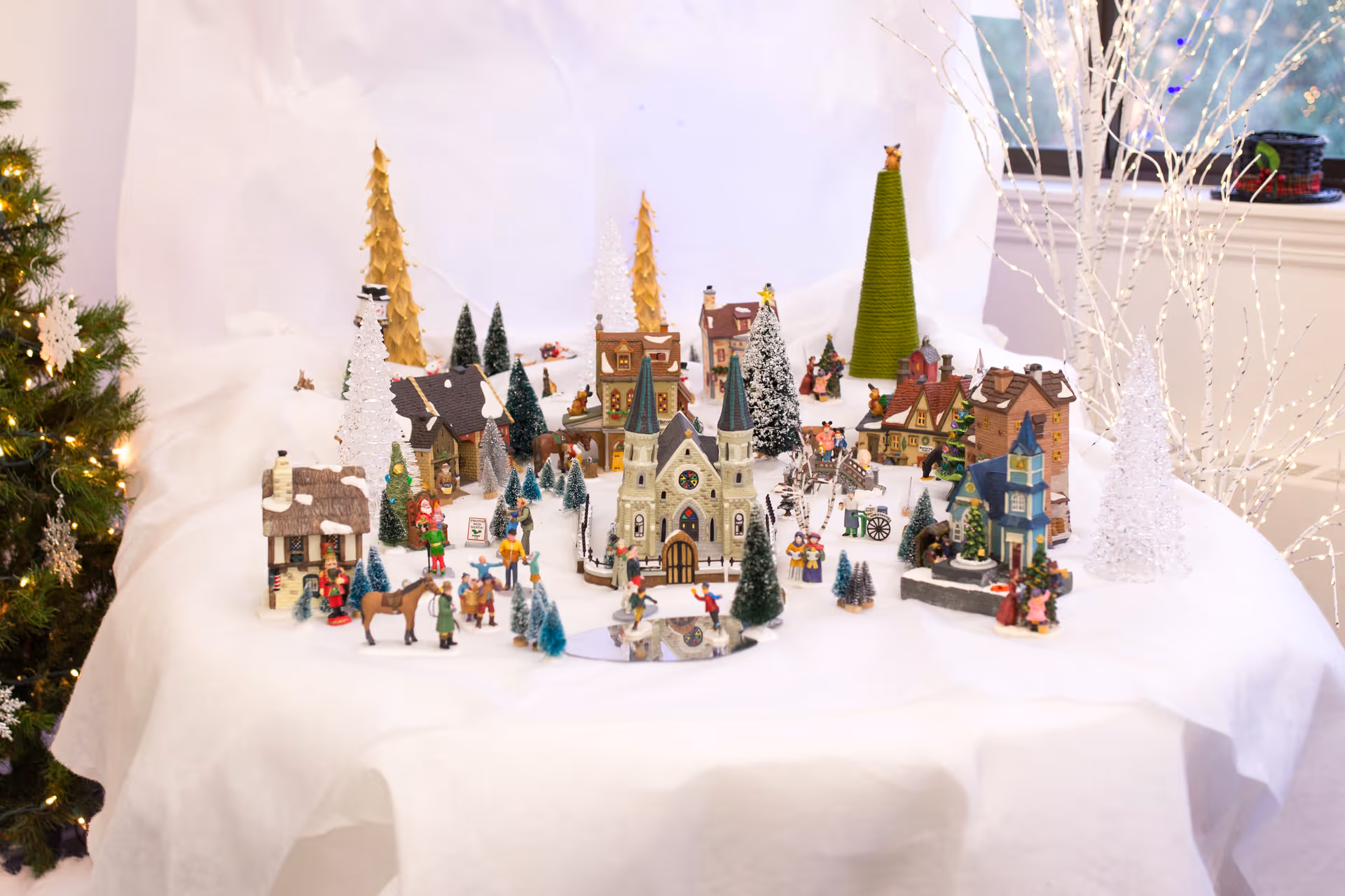 A tabletop miniature Christmas village display with tiny houses, trees, and figurines arranged on a white cloth.