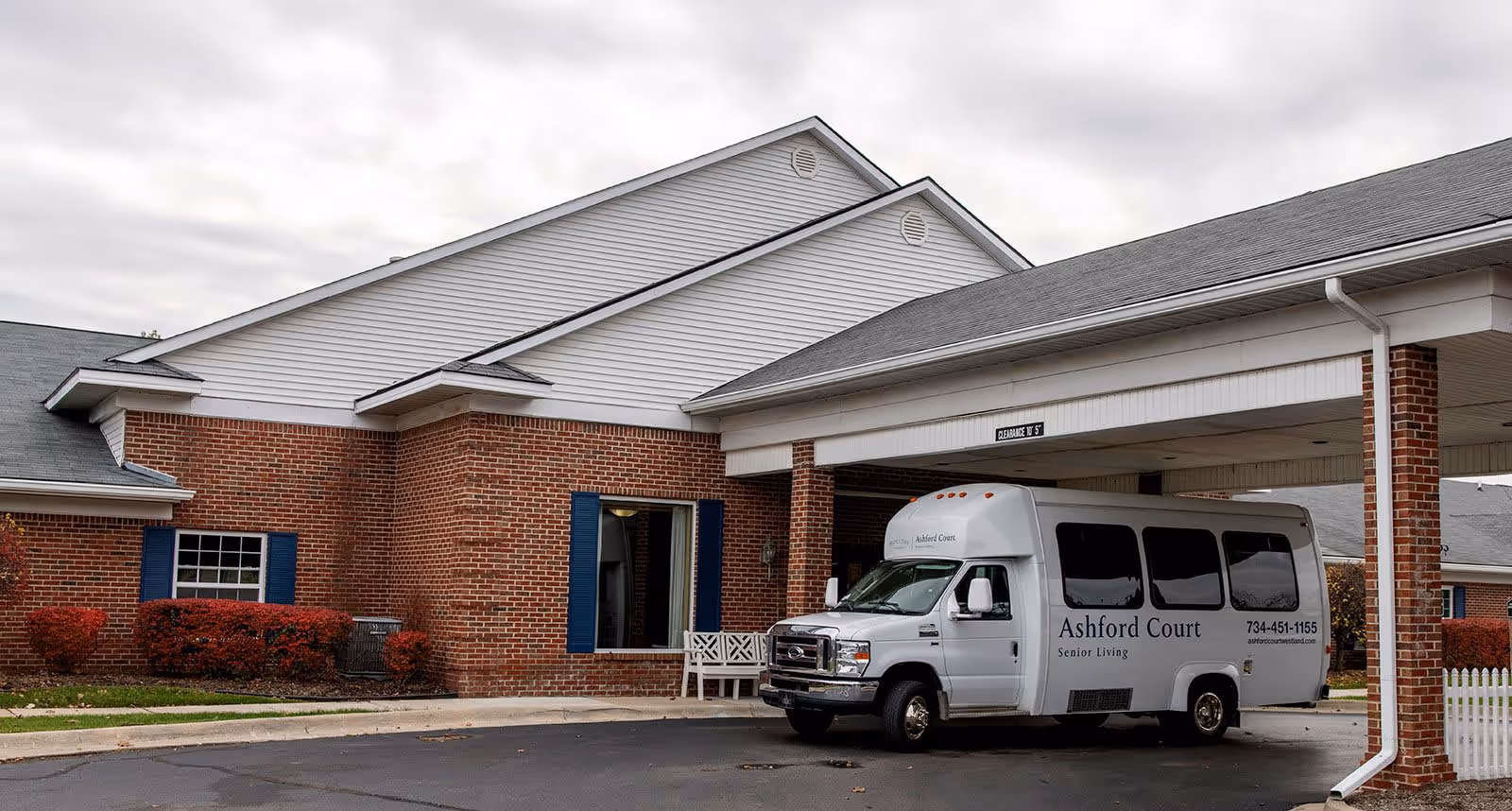 Exterior view of a senior living facility with a brick and white siding building. A white shuttle van labeled 'Ashford Court Senior Living' is parked under a covered entrance. The sky is overcast and there are some red bushes near the building.