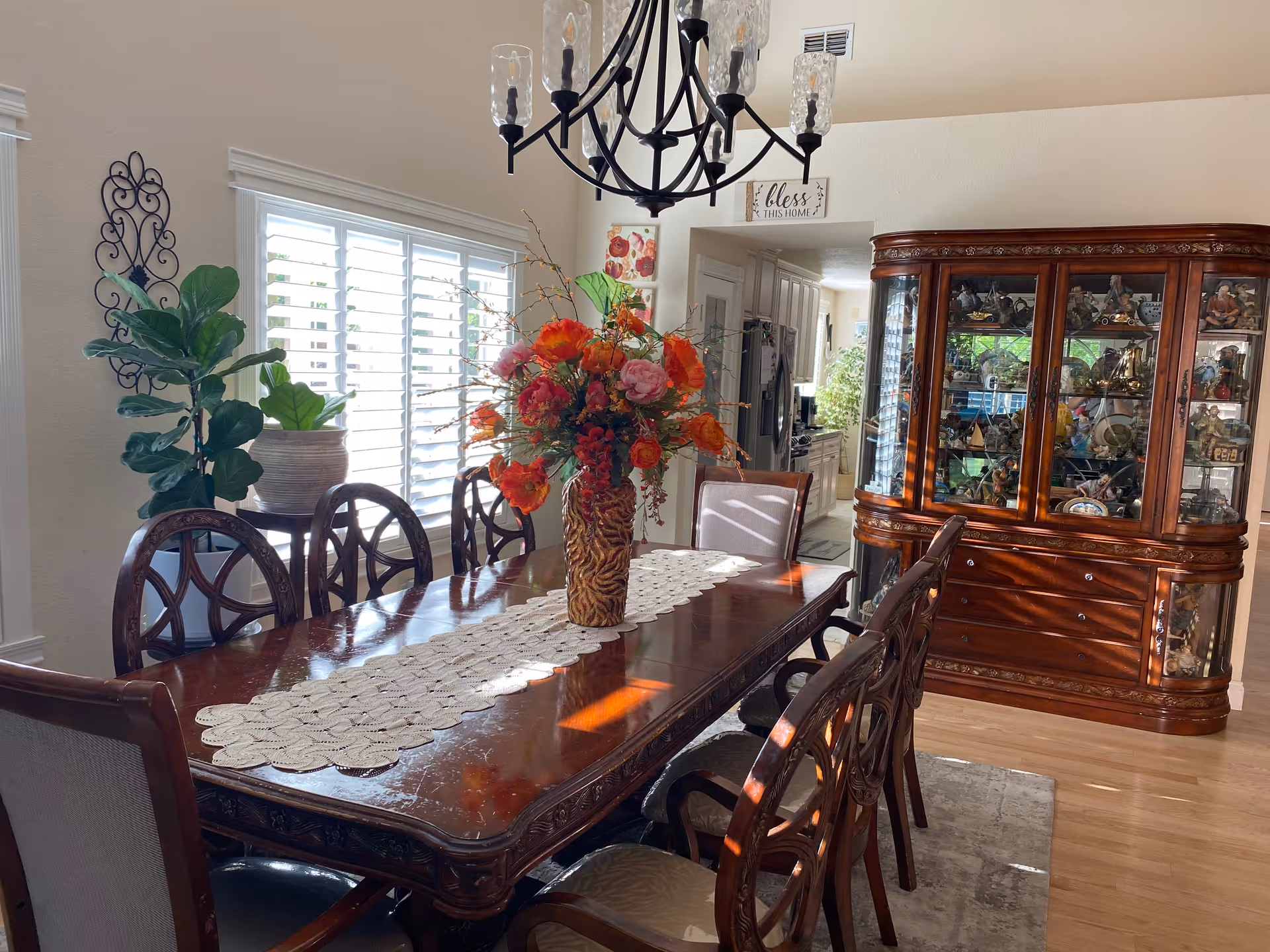A well-lit dining room with a large wooden dining table surrounded by eight chairs. The table has a lace runner and a vase with bright orange and pink flowers. There is a large wooden china cabinet filled with decorative items against the wall. A chandelier hangs above the table, and there are two potted plants near a window with white shutters. The room has light-colored walls and wooden flooring.