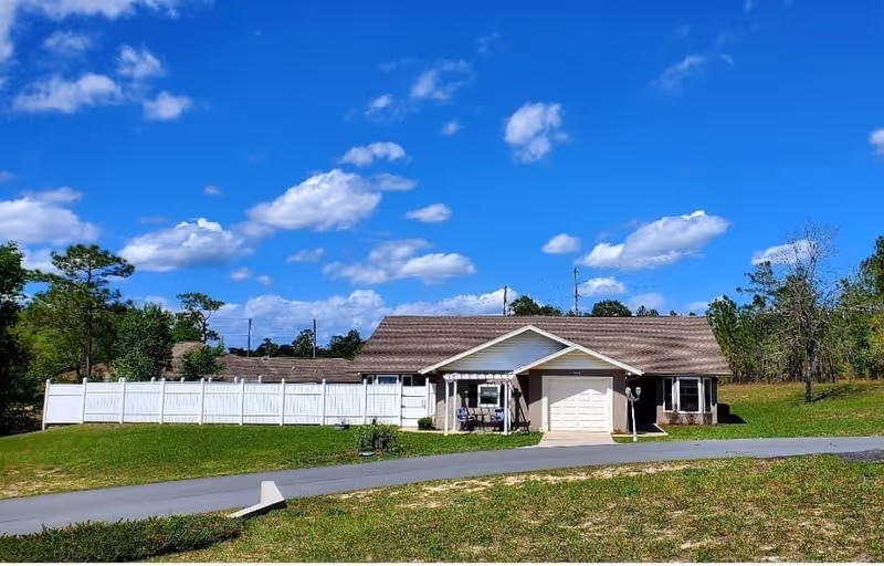 Single-story building with a brown shingled roof and a white garage door, surrounded by green grass and trees under a blue sky with scattered clouds. A white fence extends from the left side of the building.