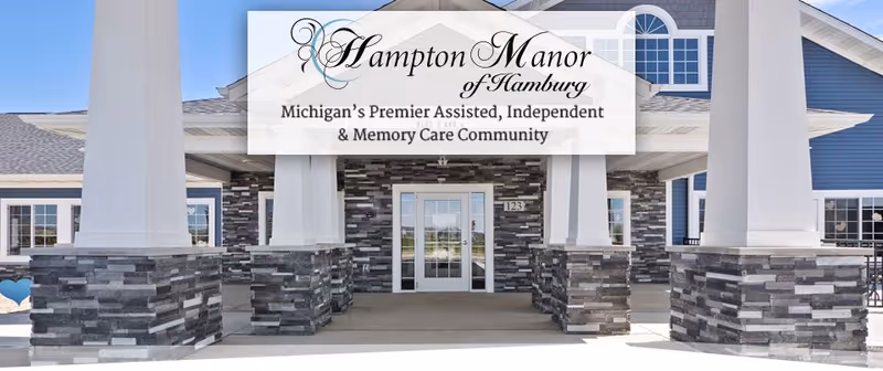 Front exterior view of Hampton Manor of Hamburg, showing the entrance with large white columns on stone bases and a covered entryway. The building features blue siding and multiple windows.