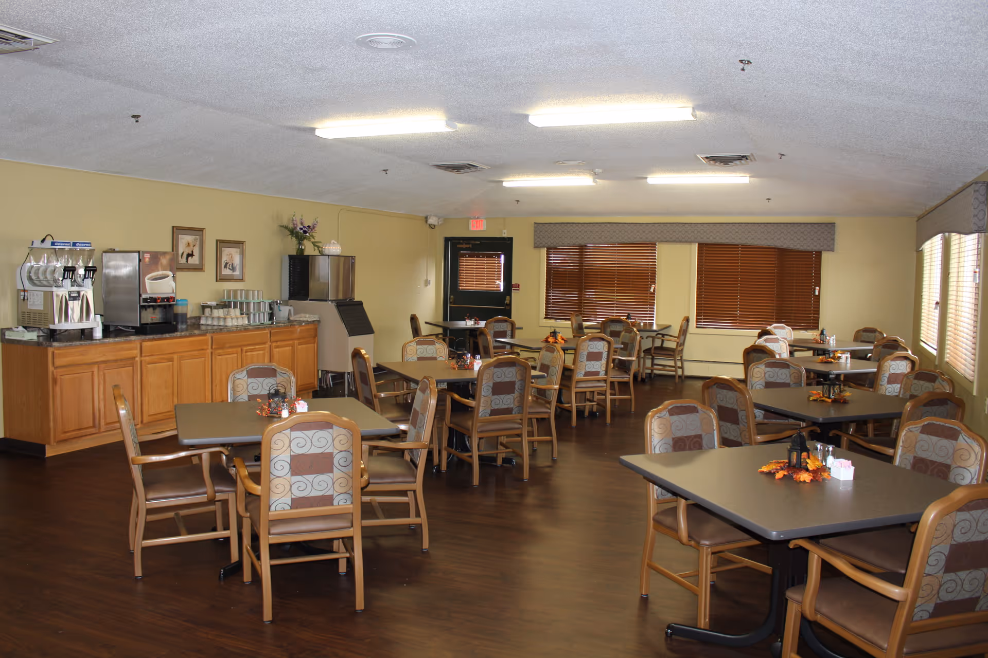 A dining room with multiple tables and chairs arranged neatly. The tables have small decorative centerpieces with autumn leaves and condiments. There is a counter along one wall with coffee and beverage machines, cups, and other supplies. The room has wooden flooring, beige walls, and windows with closed blinds.
