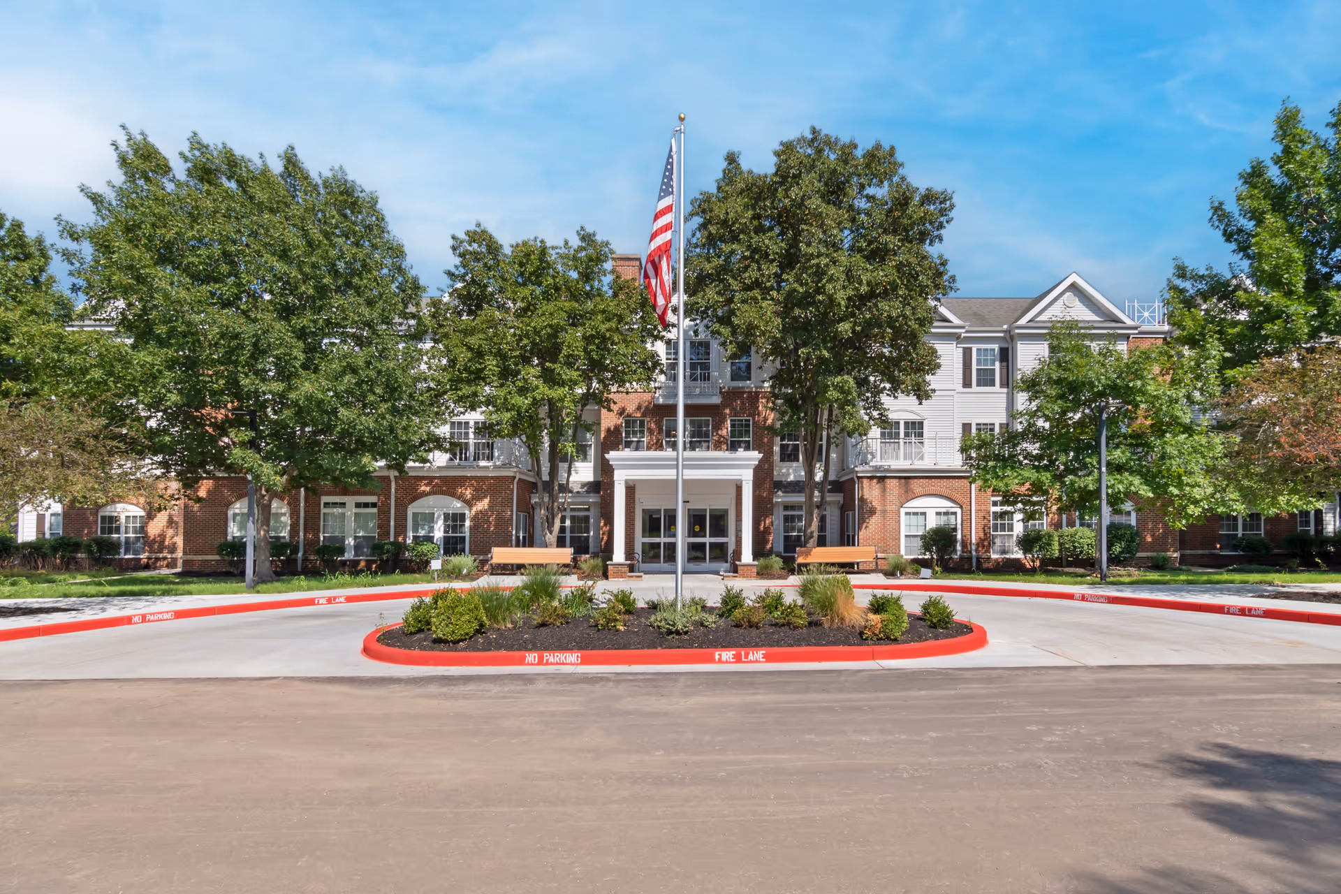 Front exterior view of a three-story senior living facility building with a circular driveway and landscaped center island featuring an American flag. The building has a mix of brick and siding with multiple windows and trees surrounding the entrance.