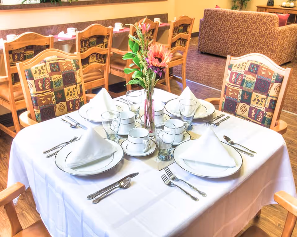 A dining table set for four with white tablecloth, white plates, folded white napkins, cups, glasses, and silverware. A vase with colorful flowers is placed in the center of the table. The background shows wooden chairs with patterned cushions and a seating area with a patterned sofa.