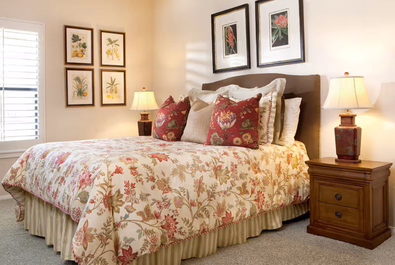 A cozy bedroom with a floral patterned bedspread and matching decorative pillows on a bed with a brown upholstered headboard. Two wooden nightstands with matching lamps flank the bed. The walls are adorned with framed botanical prints, and a window with white shutters allows natural light into the room.