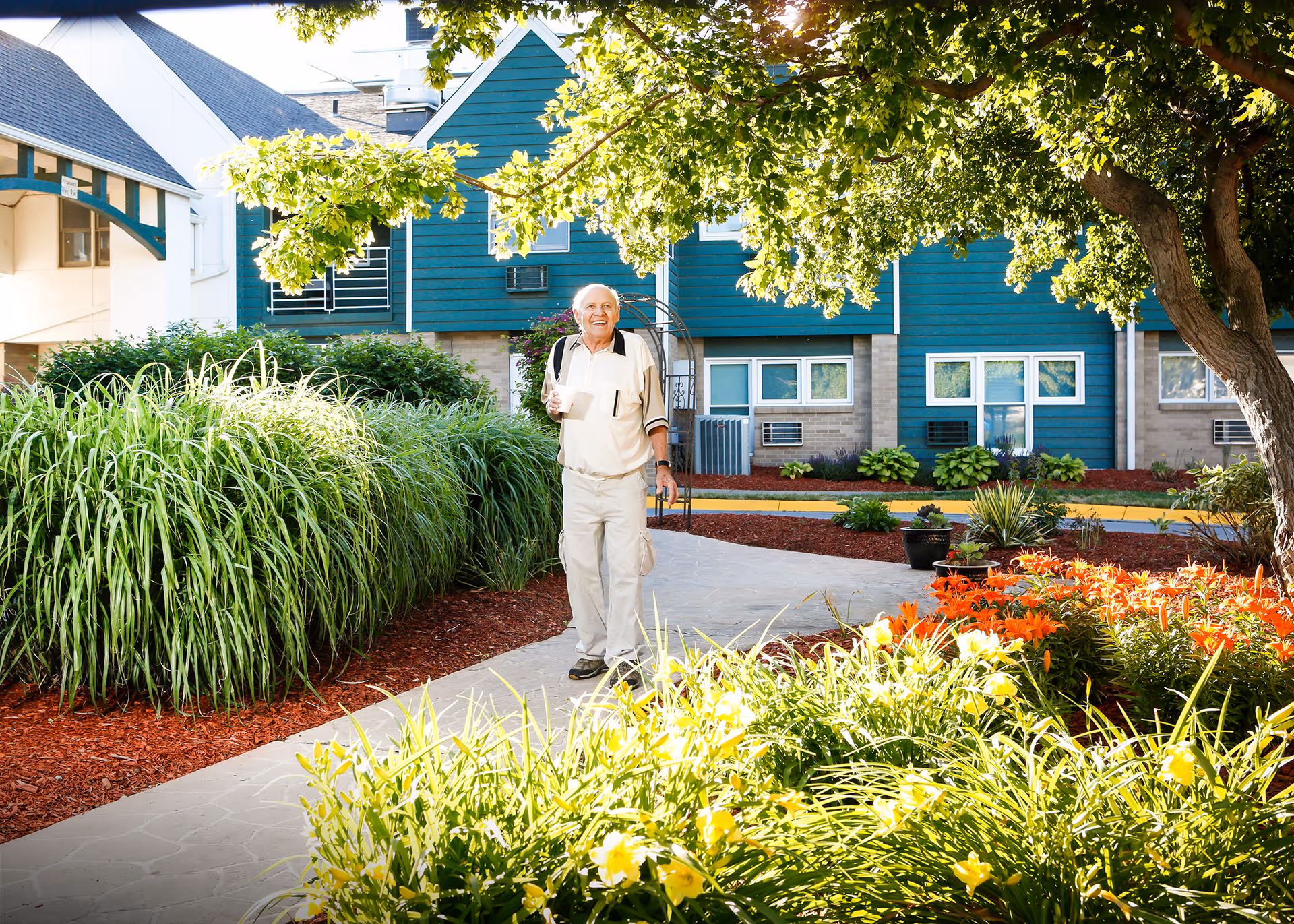 An elderly man walking along a paved pathway in a garden area with lush green plants and colorful flowers. Behind him is a building with blue siding and multiple windows. The scene is bright and sunny with trees providing some shade.