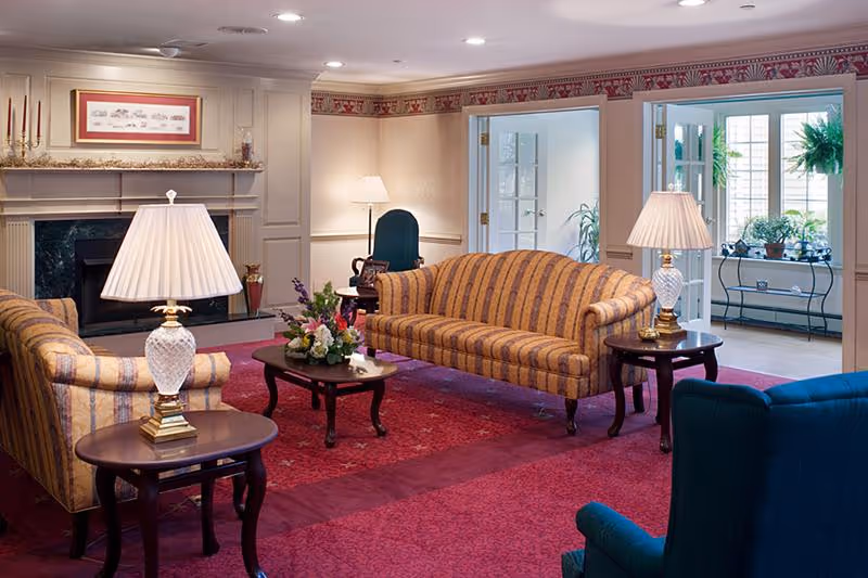 A cozy living room with striped upholstered sofas and armchairs arranged around a wooden coffee table with a floral centerpiece. The room features a red carpet, two table lamps with white shades on side tables, a fireplace with decorative items on the mantel, and a doorway leading to a sunlit room with plants on a metal table.