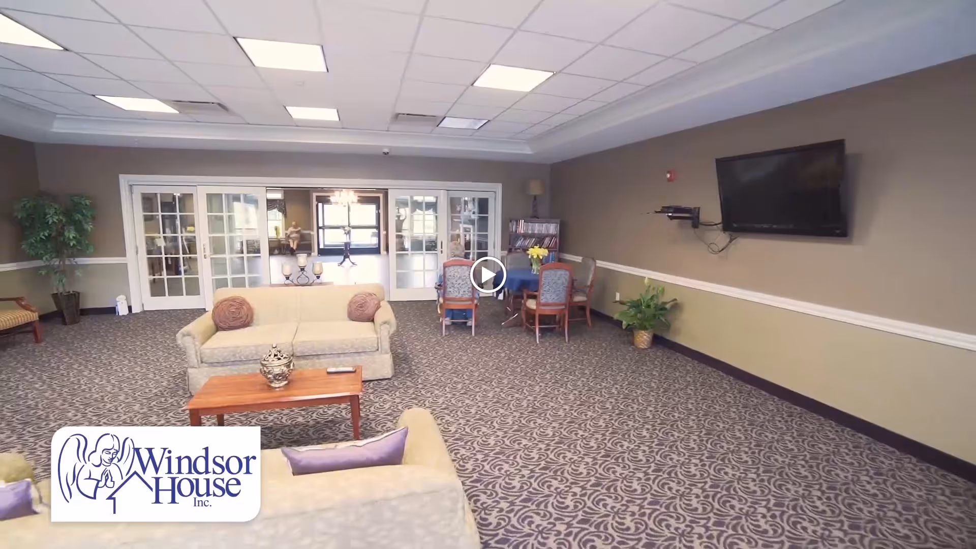 A spacious living room area with patterned carpet, beige sofas with cushions, a wooden coffee table, a wall-mounted flat screen TV, a round table with chairs, potted plants, and glass double doors leading to another room.
