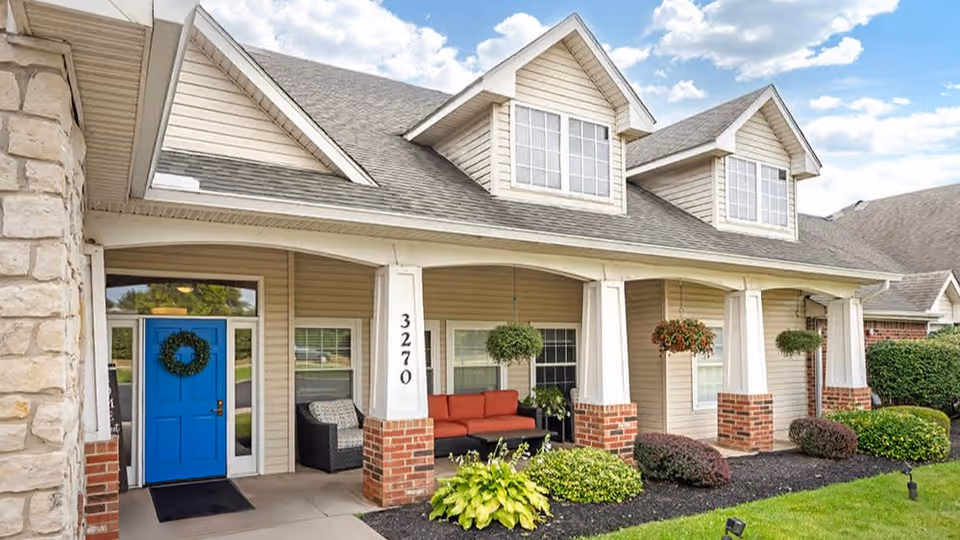 Exterior view of a residential-style building with beige siding, a blue front door decorated with a wreath, large windows, and a covered porch featuring red cushioned seating and hanging plants. The building number 3270 is displayed on a white column with brick accents. The surrounding area includes neatly trimmed bushes and a green lawn under a partly cloudy sky.
