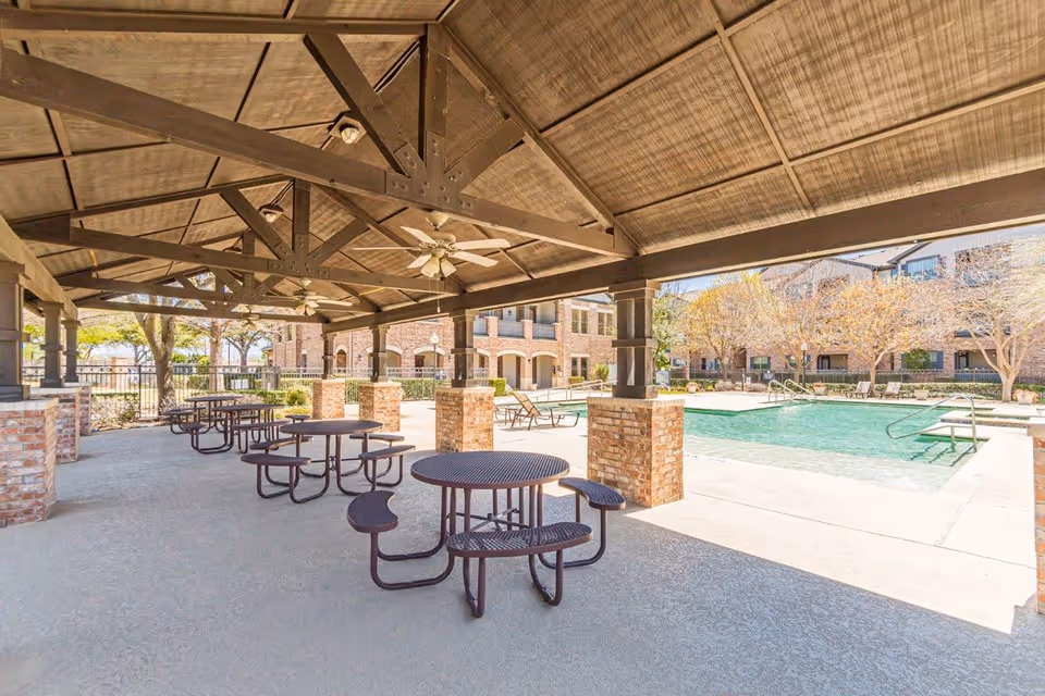 Covered outdoor patio with round metal picnic tables next to a swimming pool and apartment buildings.