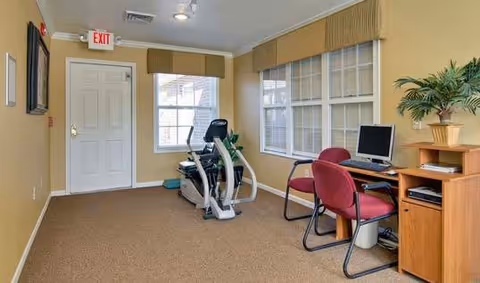 A small exercise room with beige walls and carpeted floor featuring an elliptical machine near two windows with valances. To the right, there is a wooden desk with a computer monitor, keyboard, and two red chairs. A potted plant sits on the desk. A white door with an exit sign above it is visible on the left wall.