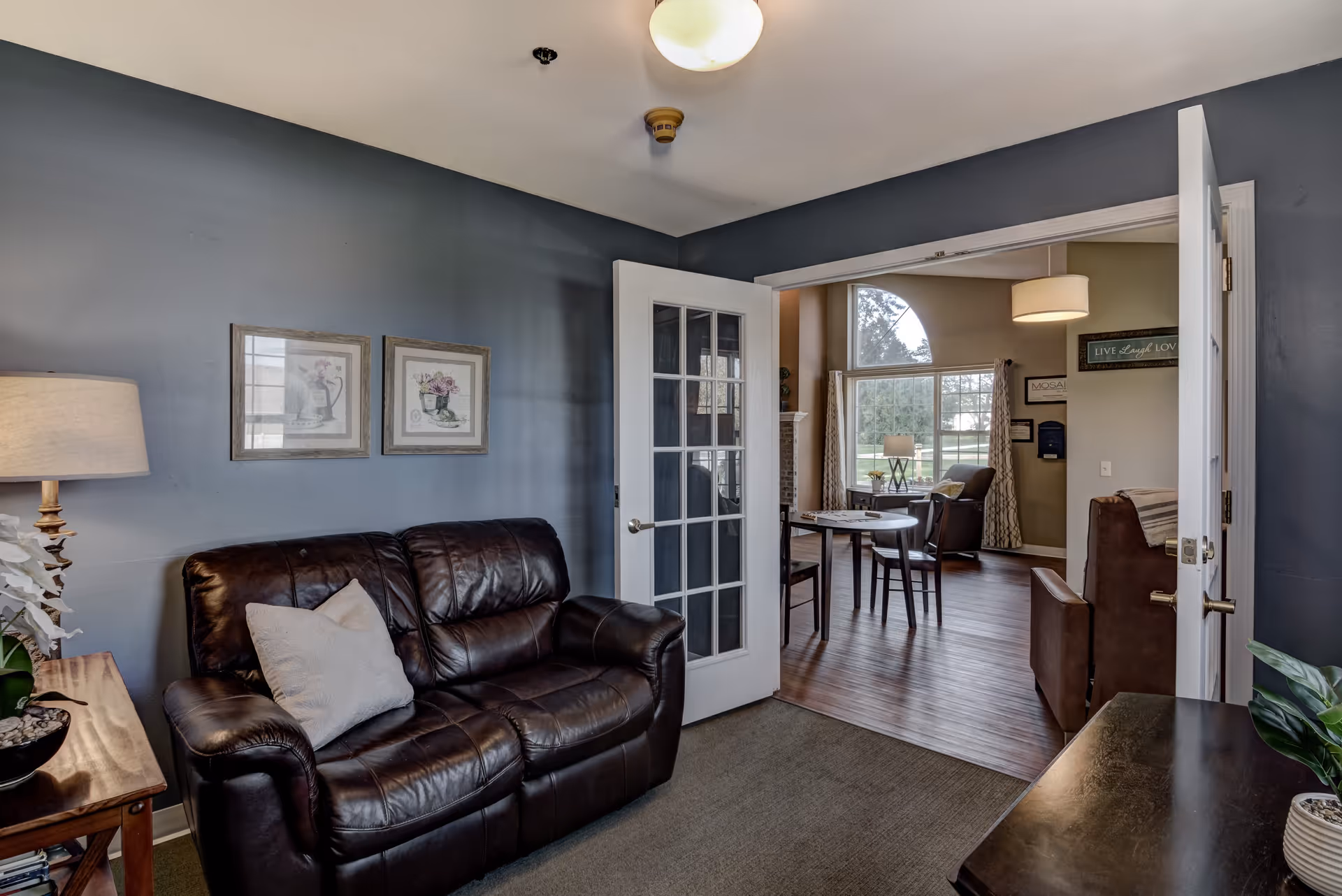 A cozy living room area with a dark brown leather loveseat, a wooden side table with a lamp and a plant, and two framed floral pictures on a blue-gray wall. Through white French doors, a dining area with a round table and chairs is visible, along with large windows letting in natural light and a view of greenery outside.