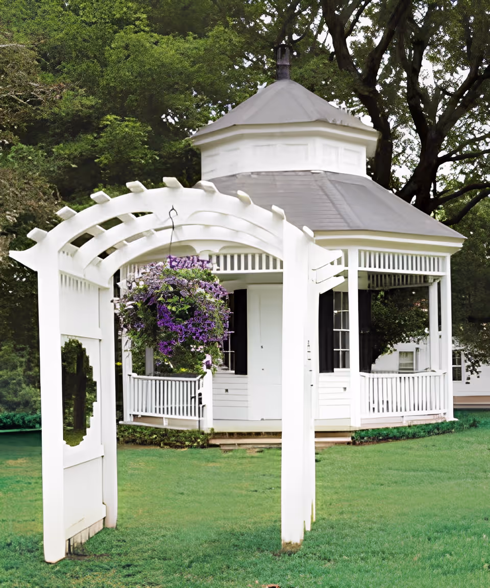 White wooden arbor leading to a white gazebo on a grassy lawn with hanging purple flowers and trees behind it.