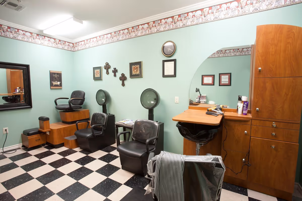 Interior of a hair salon room with black and white checkered floor, light green walls, and a decorative border near the ceiling. The room contains salon chairs, hair dryers, a wooden cabinet with a mirror, and various hair care products. Wall decorations include framed pictures and crosses.