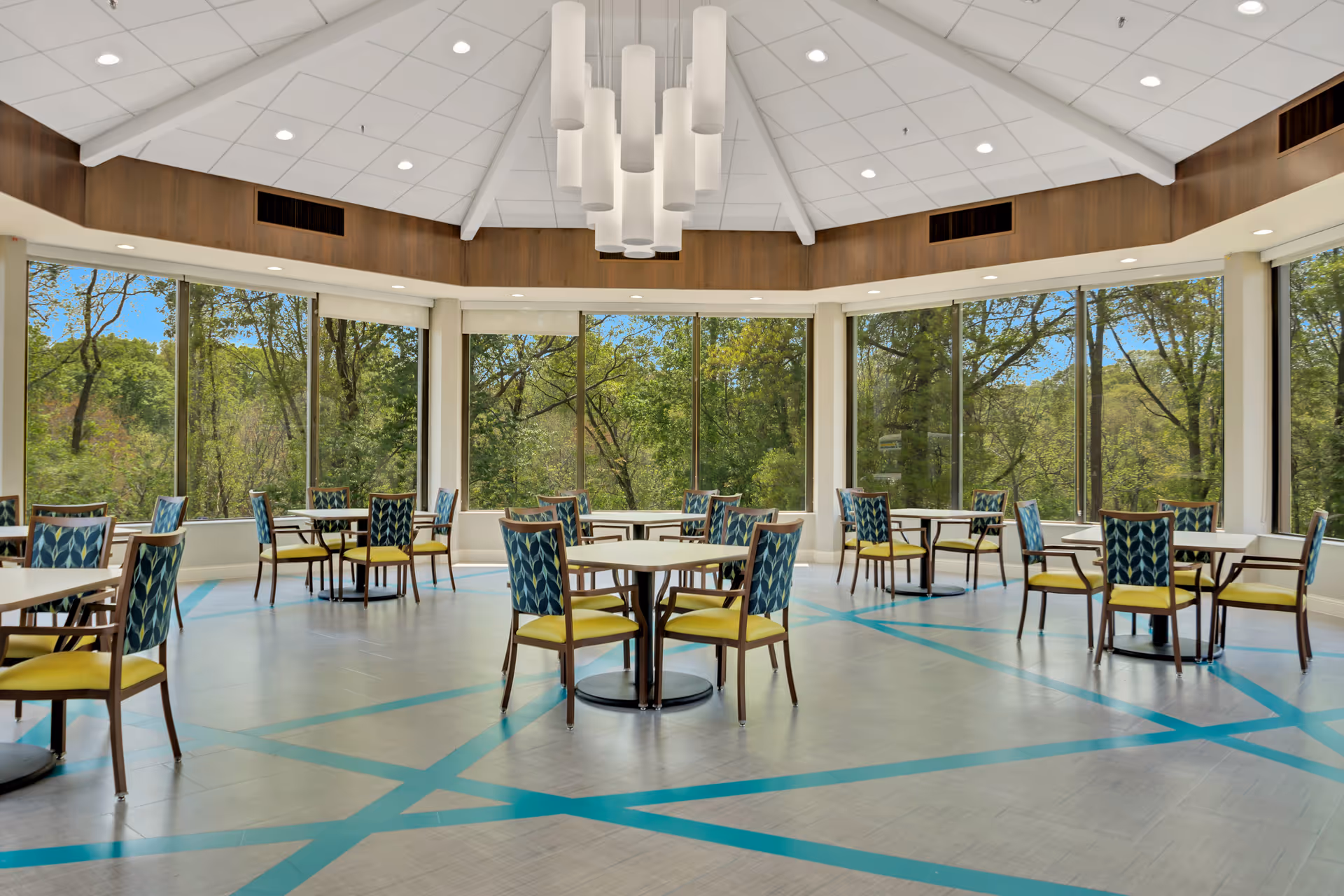 A bright dining room with multiple tables and chairs arranged neatly. The chairs have blue patterned backs and yellow seats. Large windows surround the room, offering a view of green trees outside. The ceiling is white with a modern chandelier hanging in the center.