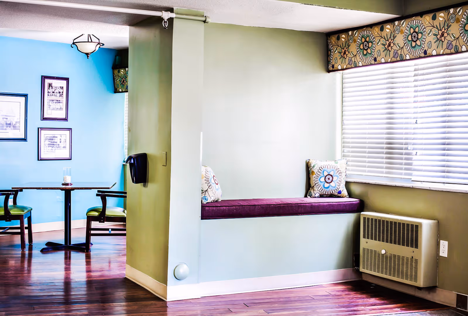 Bright common room with a purple window seat and floral pillows, a small dining table with chairs, and hardwood floors.