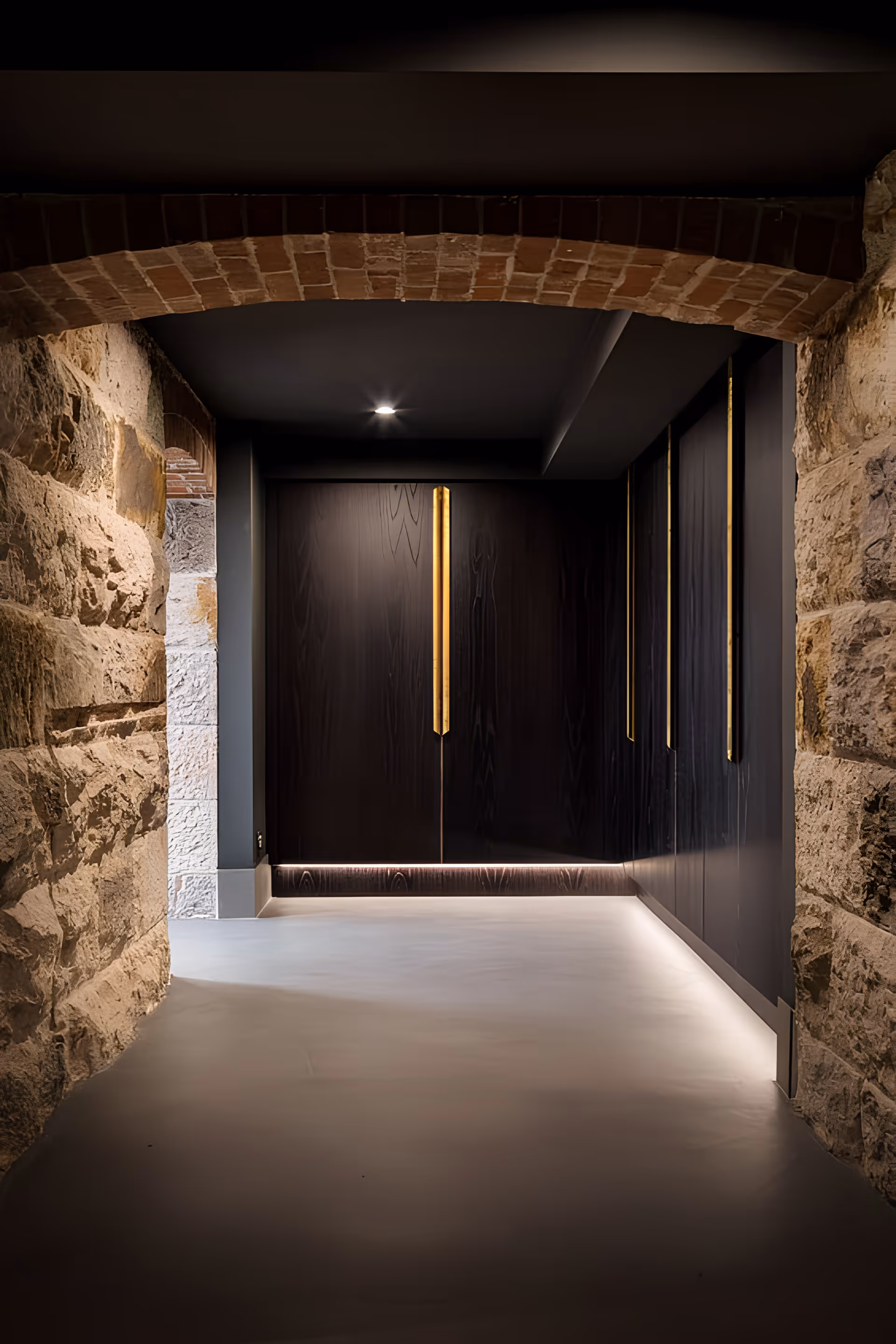 Stone-arched hallway leading to a modern dark wood cabinet wall with vertical brass handles and subtle underlighting.
