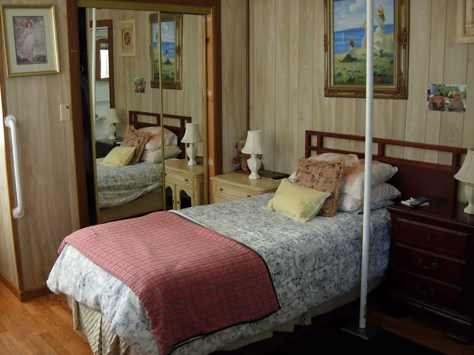 A cozy bedroom with a single bed covered in floral and patterned bedding, a red plaid blanket folded at the foot. The bed has multiple pillows and is flanked by two nightstands with white lamps. The room has wood-paneled walls adorned with framed paintings and photographs. A mirrored closet door reflects part of the room.