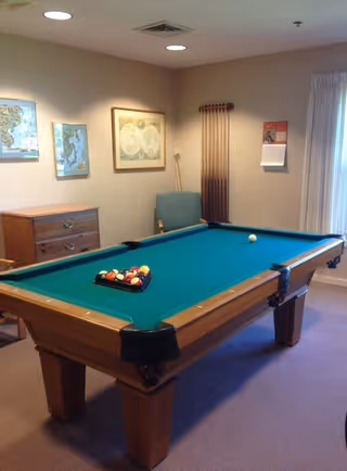A room with a pool table set up for a game, featuring a green felt surface and a rack of billiard balls. The room has beige walls with framed maps and a calendar hanging on them. There is a wooden chest of drawers, a blue chair, and a rack holding pool cues mounted on the wall. A window with white curtains allows natural light into the room.