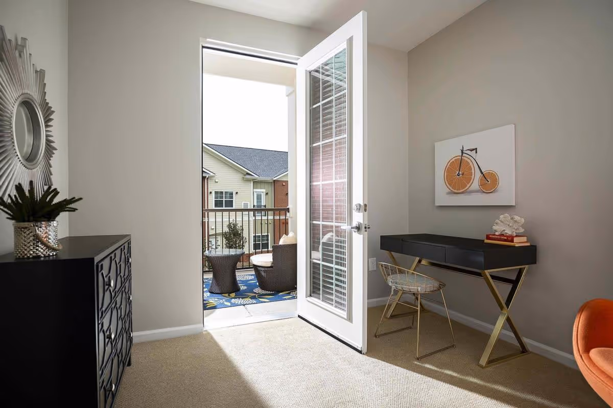 A bright room with beige walls and carpet featuring a black decorative dresser with a plant on top, a black and gold desk with a wire chair, and a wall art of a bicycle with orange wheels. A glass door is open leading to a balcony with outdoor wicker furniture and a blue patterned rug. A neighboring building is visible outside.