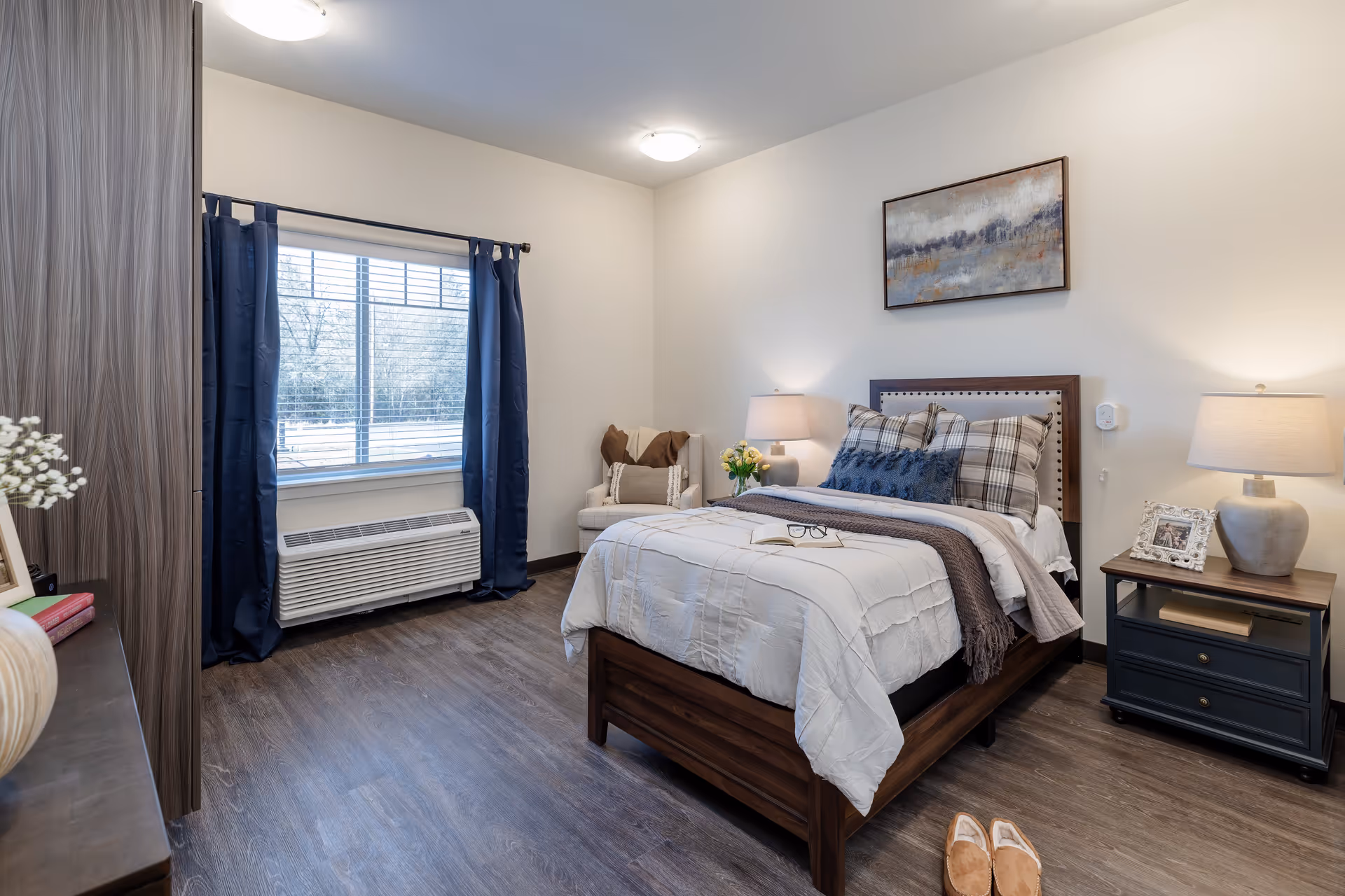 A cozy bedroom in a memory care facility featuring a wooden bed with white and plaid bedding, a nightstand with a lamp and framed photo, a cushioned chair with a throw blanket, a window with navy blue curtains, and wood flooring.