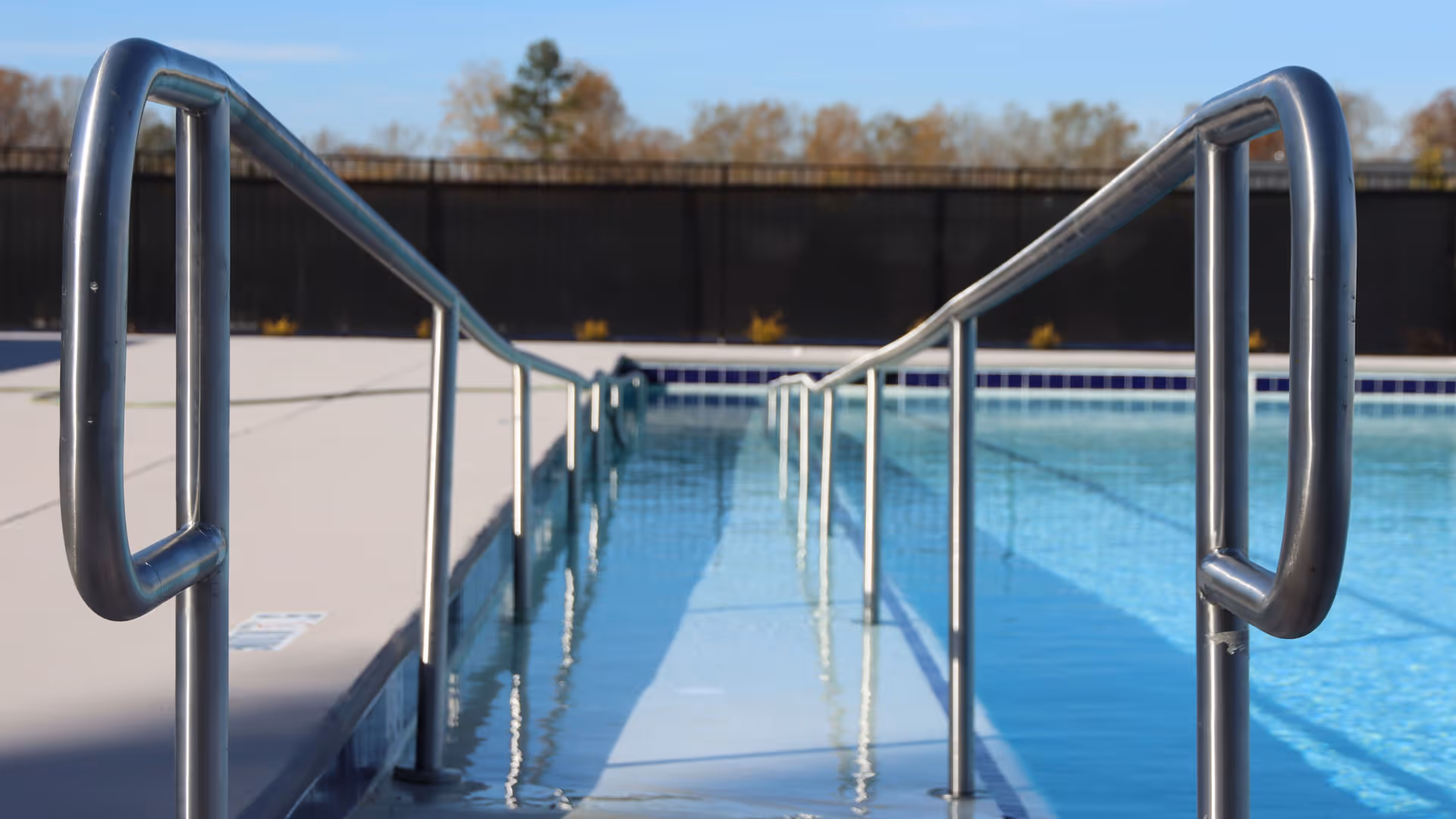 A close-up view of a swimming pool ramp with stainless steel handrails on both sides, leading into clear blue water. The pool deck is visible on the left side, and a black fence and trees are in the background under a clear sky.