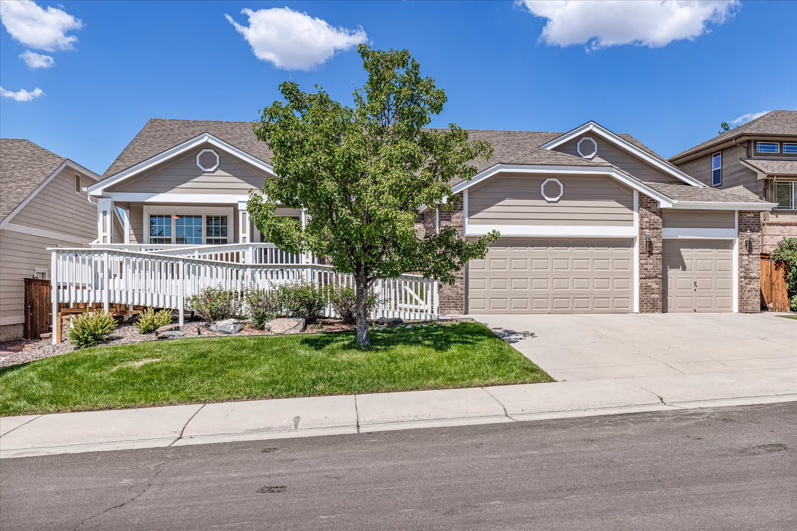 Exterior view of a single-story residential building with a beige facade, a large garage with three doors, a white wheelchair-accessible ramp leading to the front entrance, a green lawn, and a tree in front under a partly cloudy blue sky.