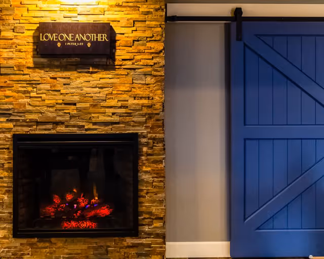 A cozy interior scene featuring a stone wall with a built-in electric fireplace emitting a warm glow. Above the fireplace is a wooden sign with the inscription 'LOVE ONE ANOTHER 1 PETER 1:22'. To the right of the fireplace is a blue sliding barn door mounted on a black metal track.