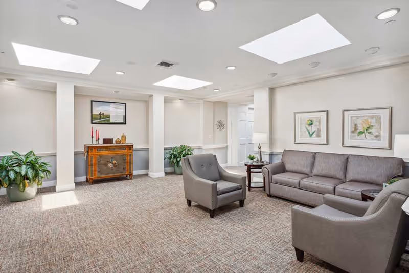 A bright and spacious living room area with beige walls and carpeted floor. The room features a gray sofa, two gray armchairs, two side tables with lamps, potted plants, framed floral artwork on the walls, and a wooden cabinet with decorative items. The ceiling has recessed lighting and skylights allowing natural light to fill the space.