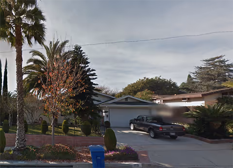 A residential street view showing a single-story house with a garage and a black pickup truck parked in the driveway. The front yard has various trees and shrubs, including palm trees and a small deciduous tree with autumn leaves. A blue recycling bin is placed near the curb.